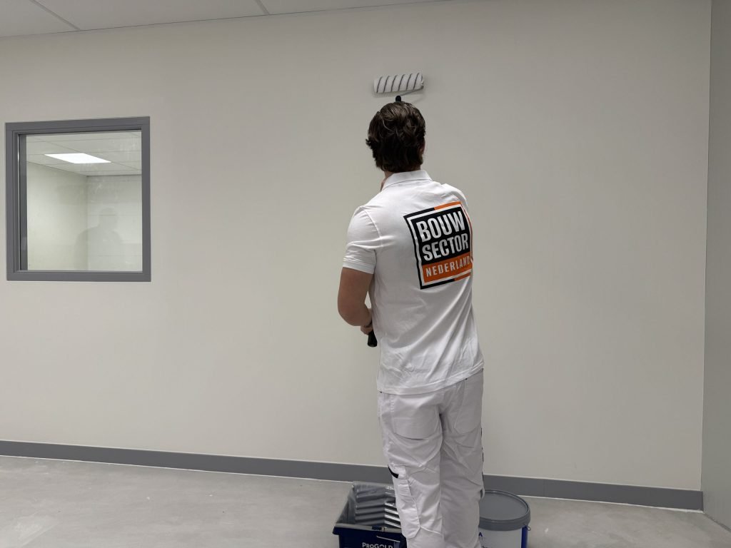 Schilder van Bouwsector Nederland rolt een lichte muur onder het plafond, met verfbak en emmer bij de plint