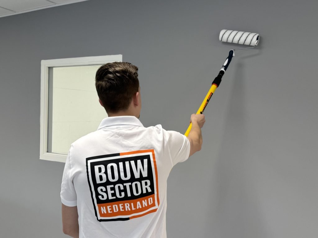 Schilder van Bouwsector Nederland zet een grijze muur in de verf met een roller op een verlengsteel
