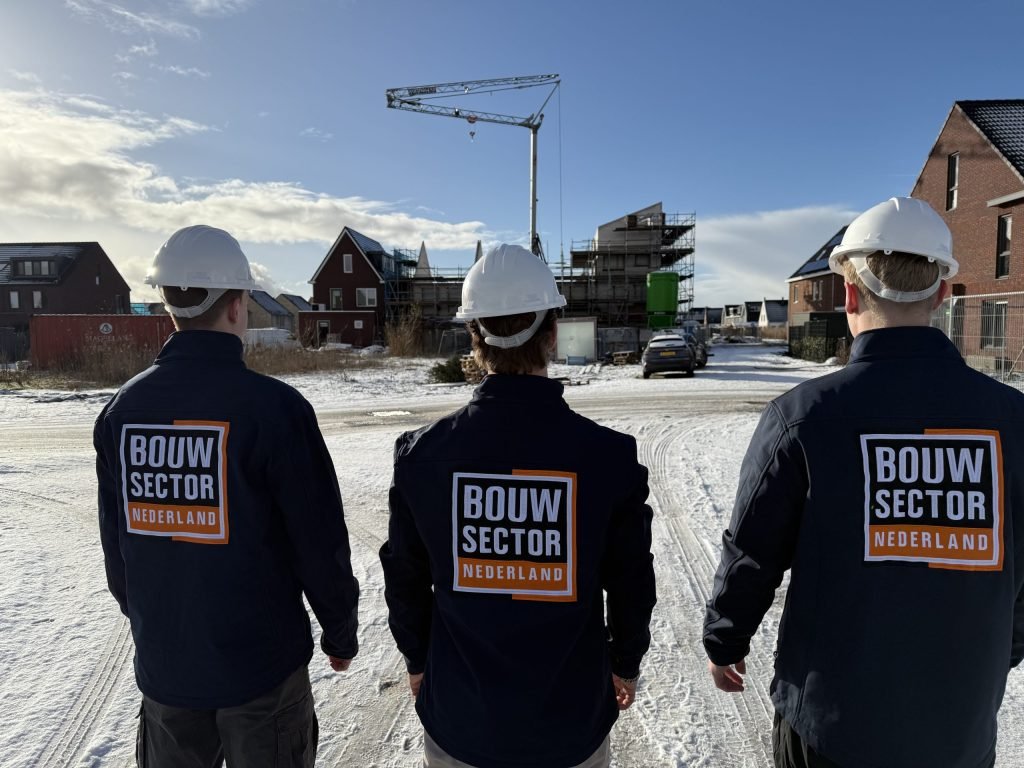 Drie vakmensen van Bouwsector Nederland met witte helm en bedrijfsjas op een besneeuwde bouwweg bij woningen in aanbouw